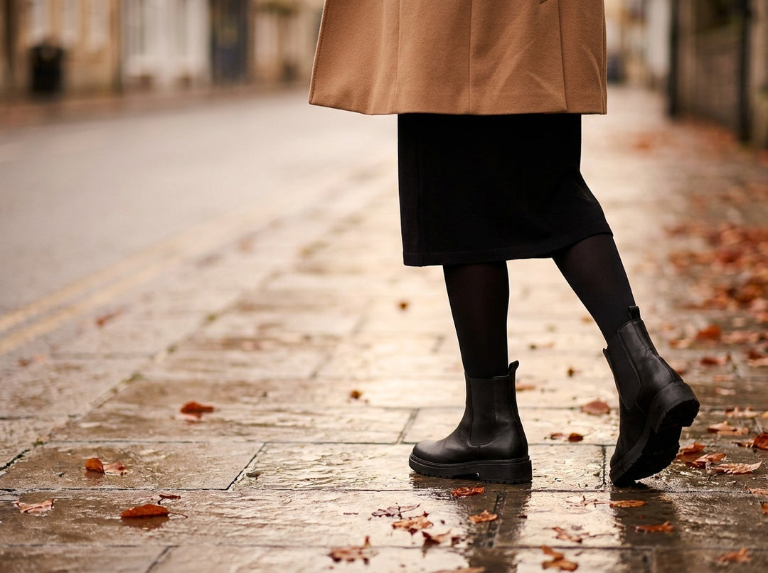 Legs in black thermal tights with a black midi skirt, the hem of a camel coat and black chunky ankle boots, walking on a wet autumn pavement scattered with fallen leaves.